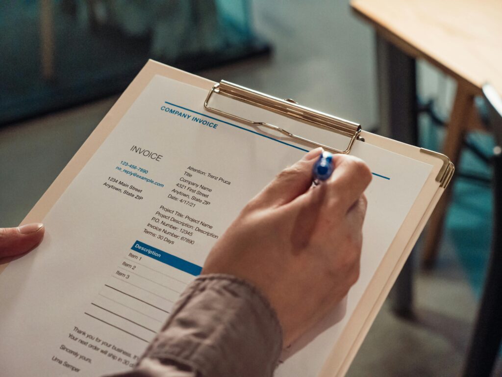 Person's hand holding a company invoice on a clipboard with a pen.