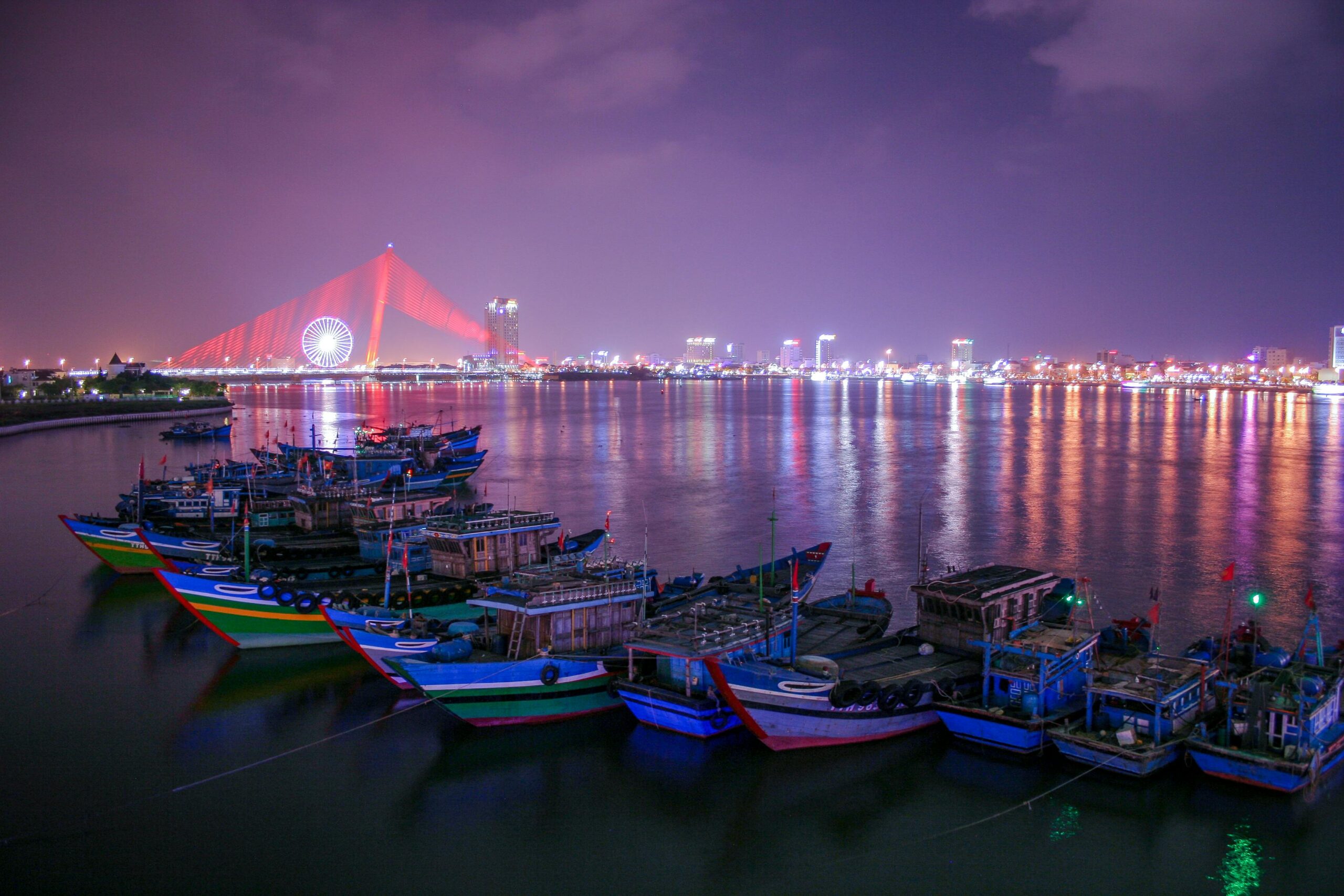 Colorful boats docked at Da Nang's waterfront with Dragon Bridge illuminated at night, offering a stunning cityscape.